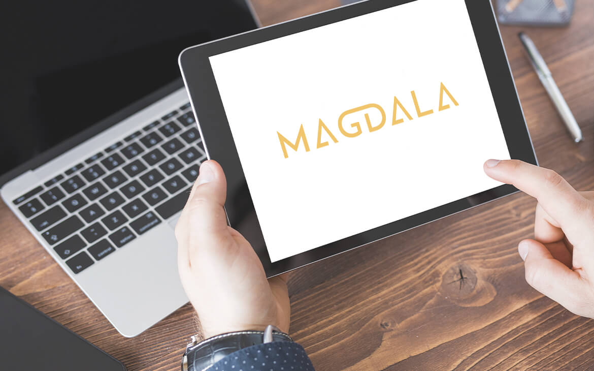 Um ipad, com notebook e na tela o logo Magdala em formato de visual aid